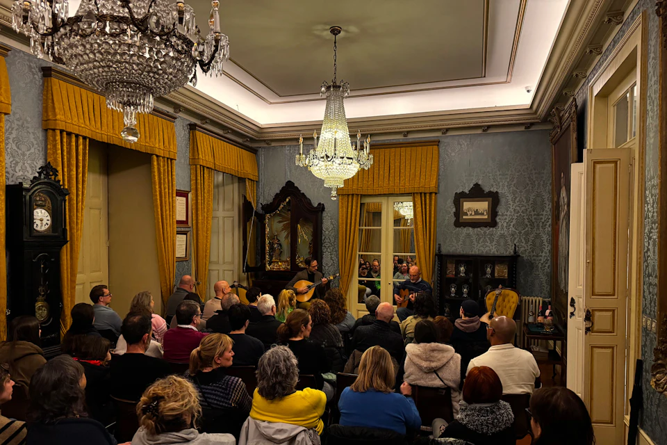 A group of people sitting in a room watching a performance under a chandelier, with ornate decor and yellow curtains.