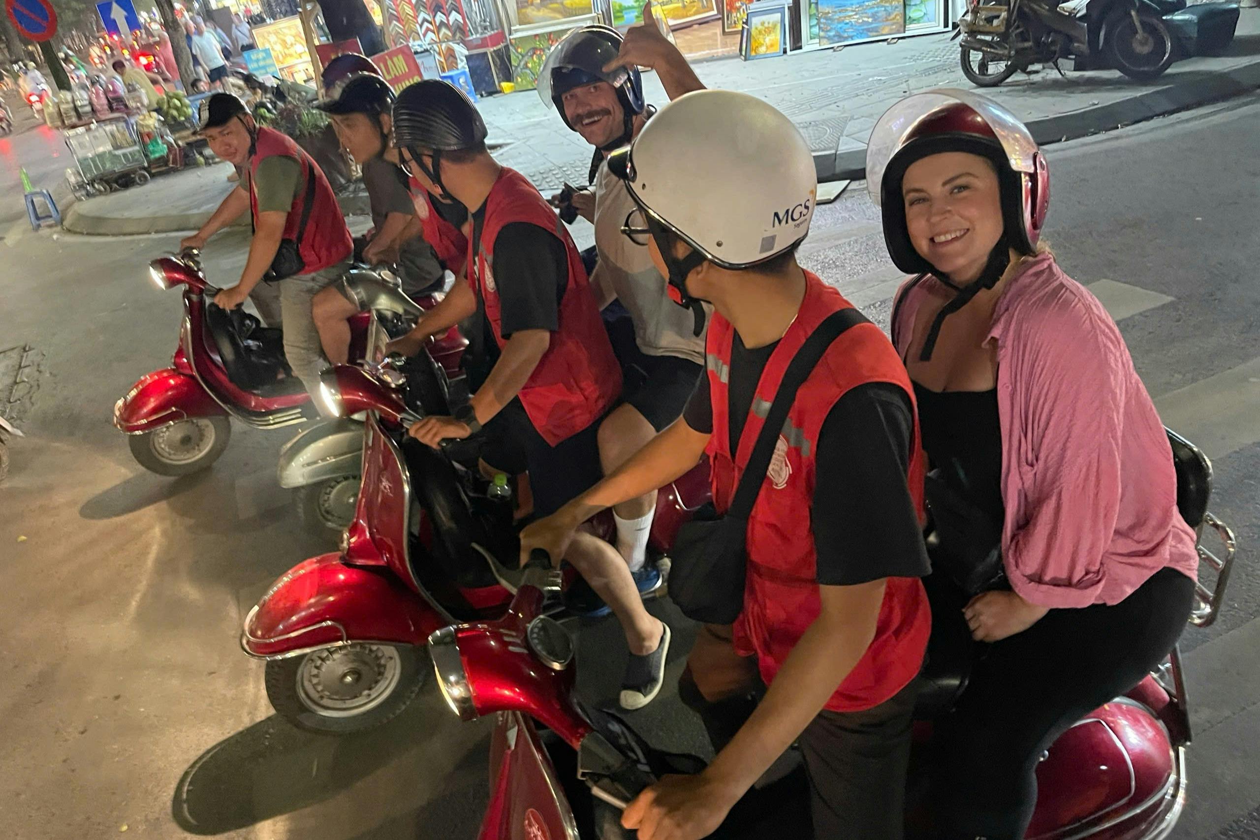 Two people wearing helmets sit on a scooter at night; the passenger, a smiling woman, is wearing a pink shirt.