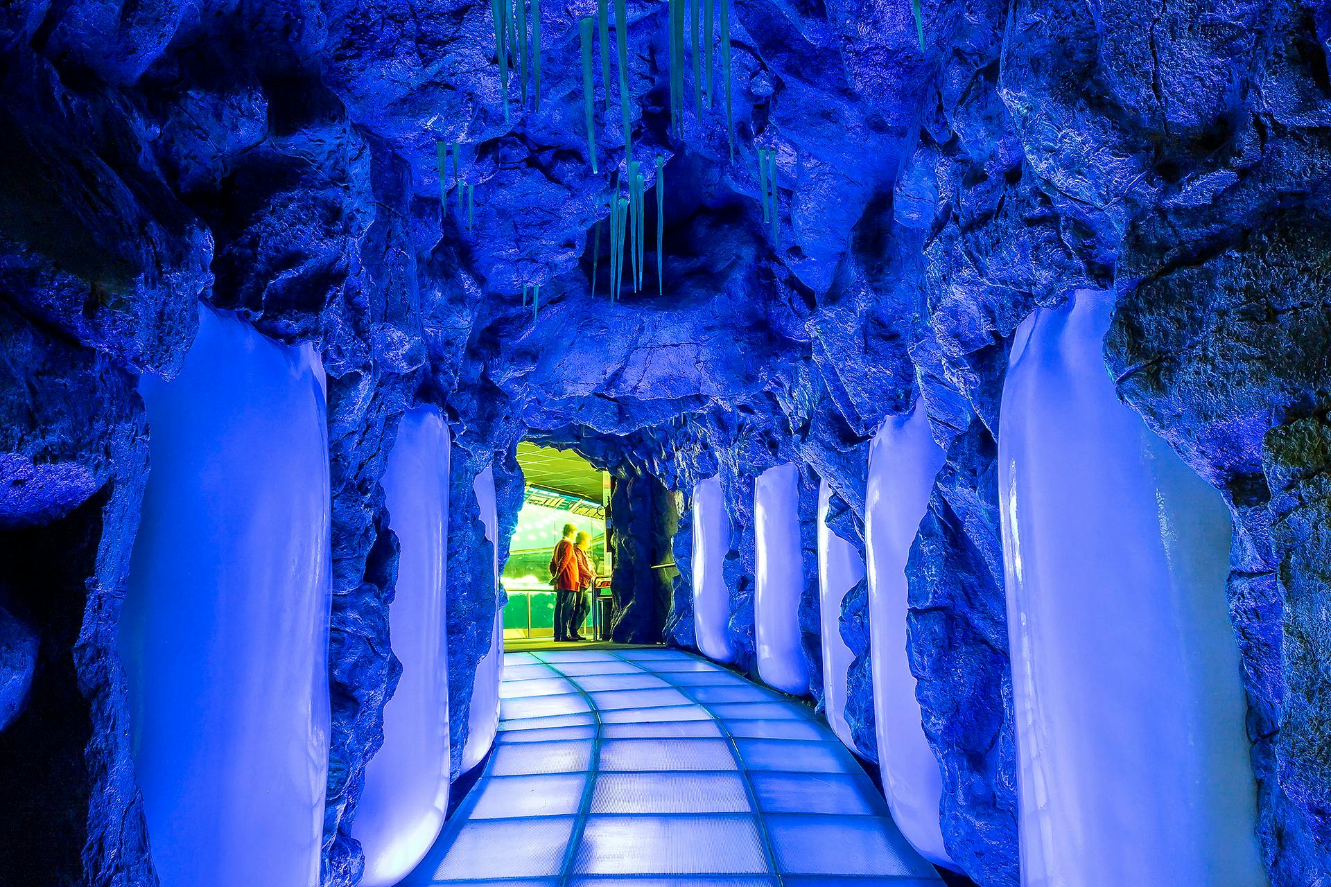 험준하고 푸른 바위 벽과 반투명한 바닥이 있는 밝은 조명의 터널 끝에 사람들이 서 있습니다.