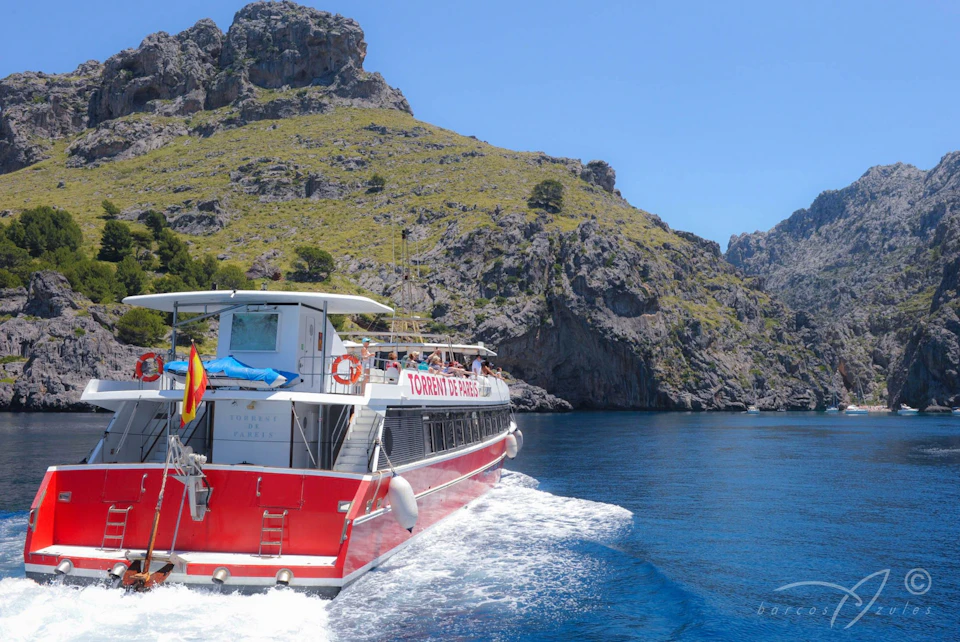 Red and white boat with "Torrent De Pareis" written on the side, cruising on blue water with rocky hills in the background.
