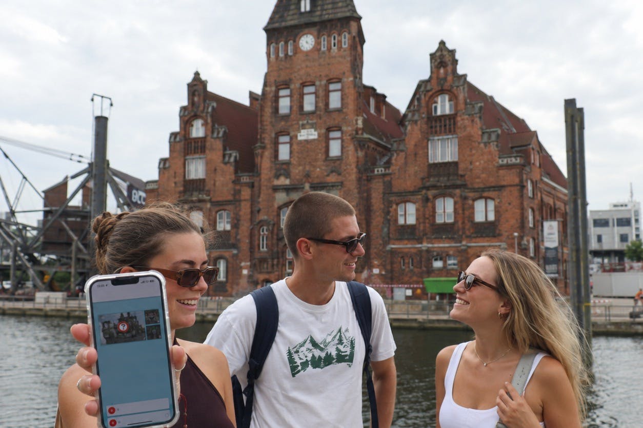 Duas pessoas a sorrir ao ar livre, perto de um edifício histórico de tijolo e de um curso de água, uma delas a segurar um smartphone com o ecrã visível.