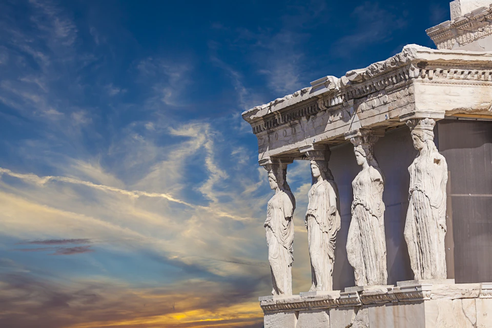 Ancient Greek temple facade with four caryatid statues standing as columns against a vibrant, sunset-lit sky.
