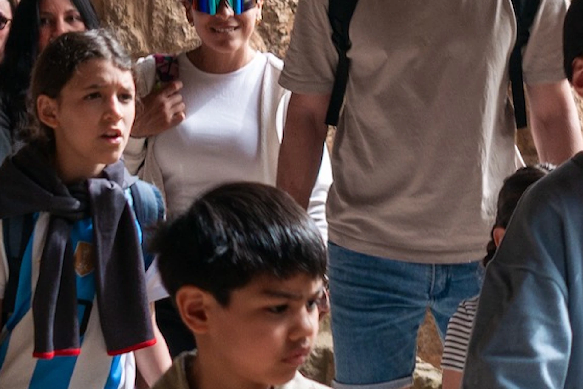 A group of people walking through an area with stone archways and sunlight filtering through. Children are holding papers.