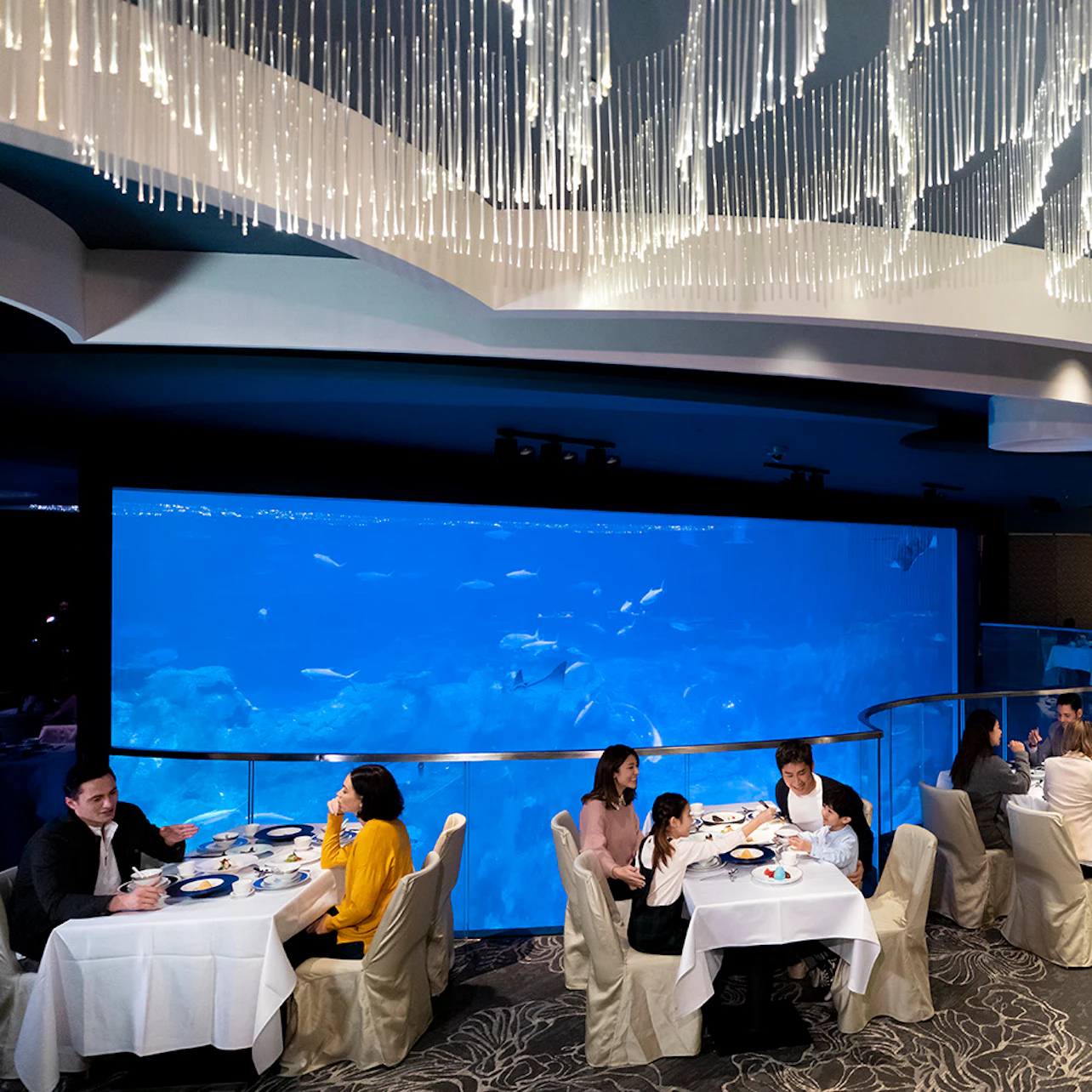 A restaurant with diners seated at tables, featuring a large aquarium wall and a decorative ceiling with hanging lights.