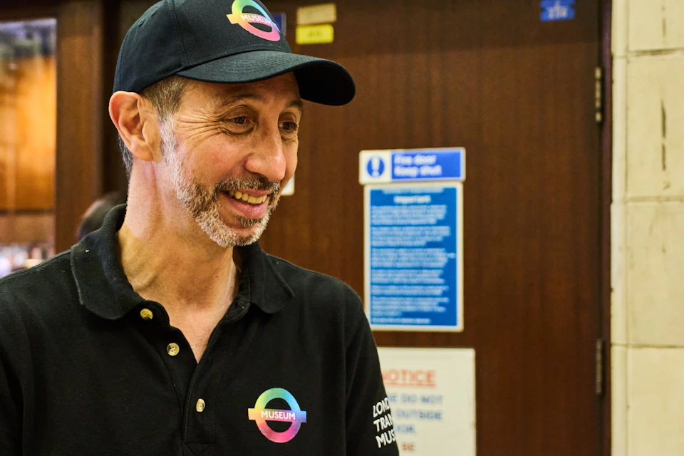 A man in a black uniform with a rainbow logo interacts with two individuals in a public indoor setting.