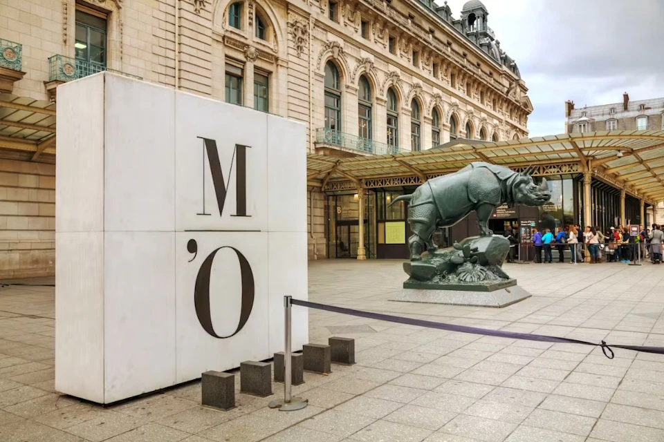 A building with arched windows, a rhinoceros statue, and a large 'M O' sign in front of a museum entrance.