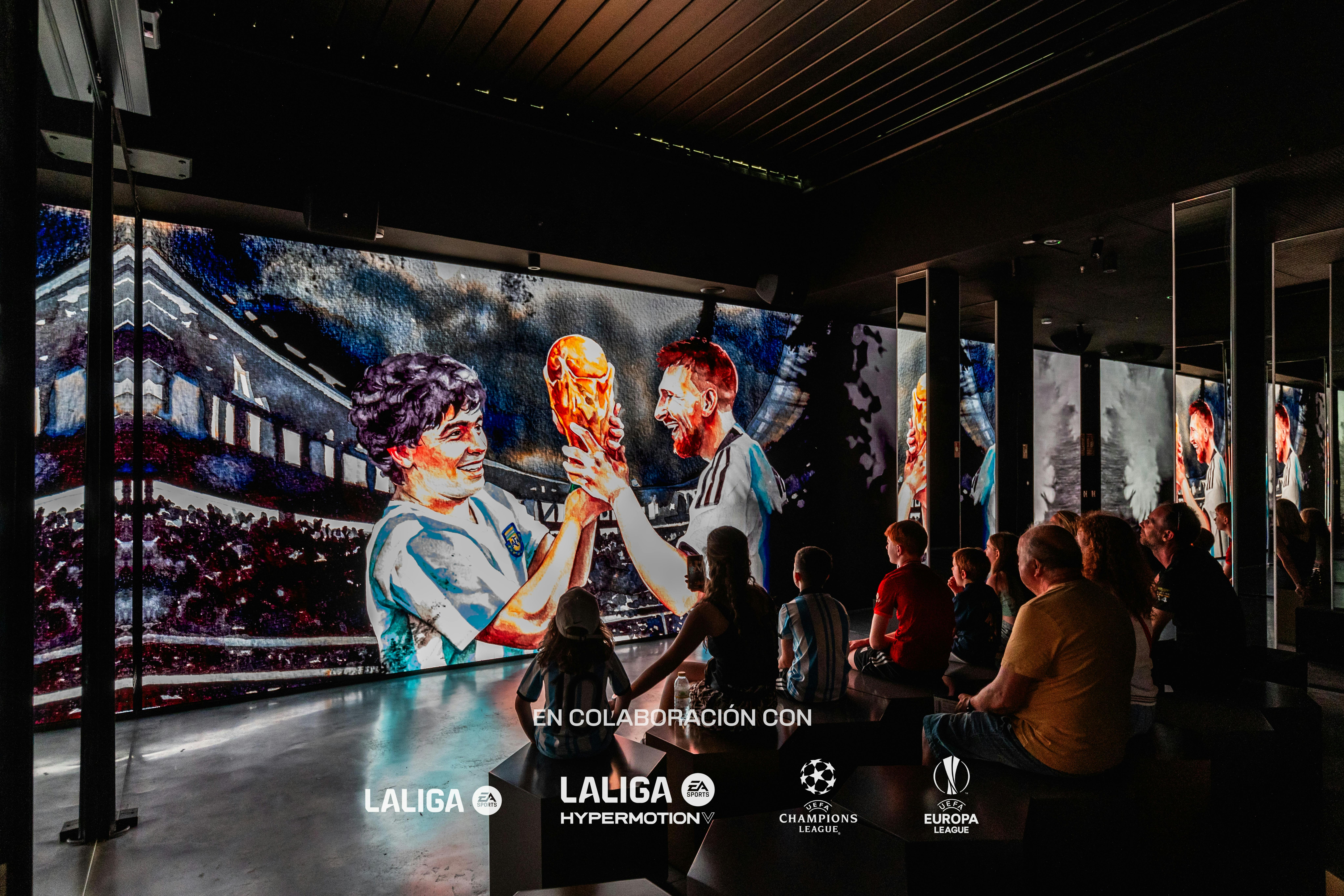 Menschen sitzen auf Bänken in einem dunklen Raum und schauen auf einen leuchtenden Bildschirm, auf dem zwei animierte Fußballspieler einen Pokal halten.