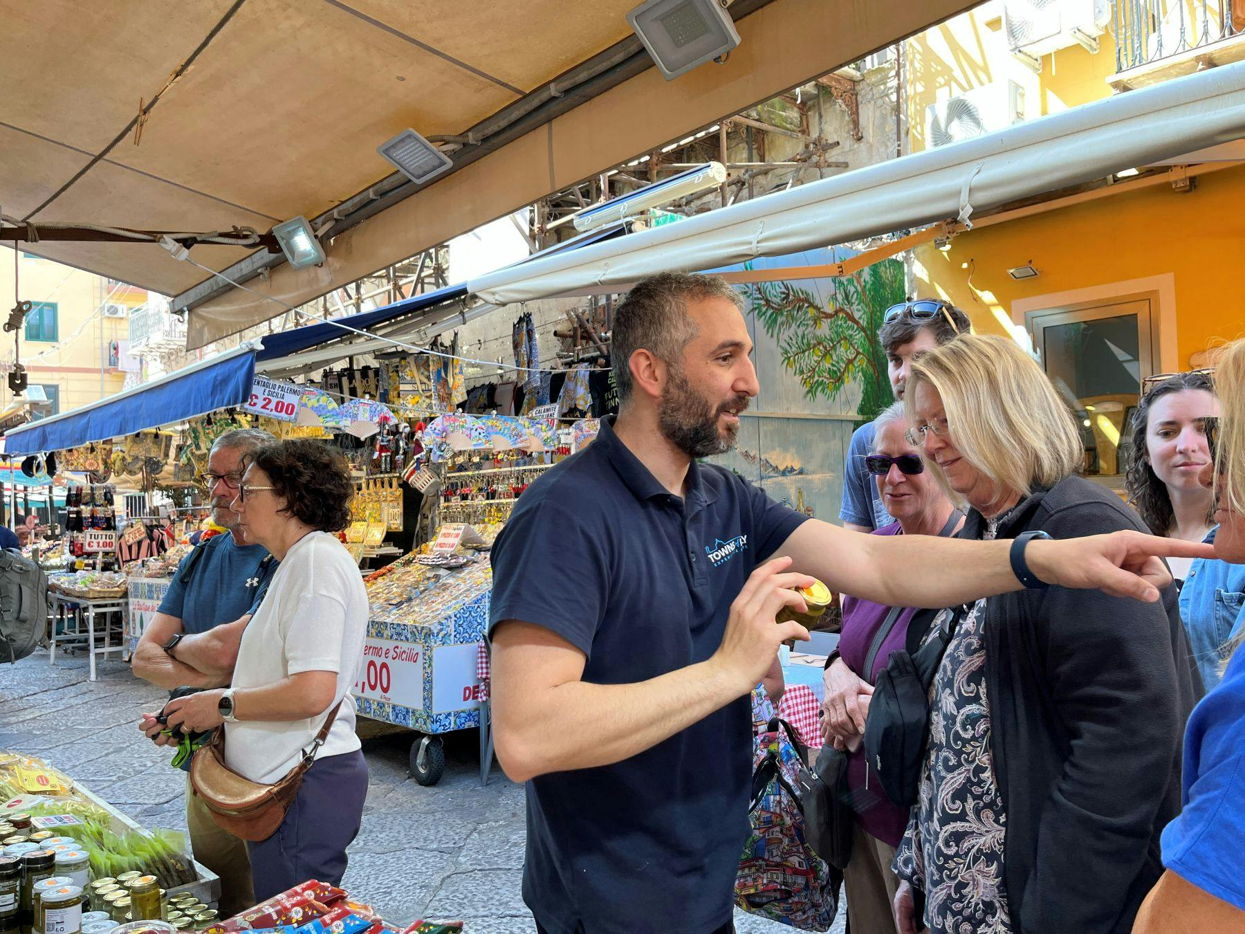 A man gestures while speaking to a group of people at an outdoor market with colorful stalls and merchandise.