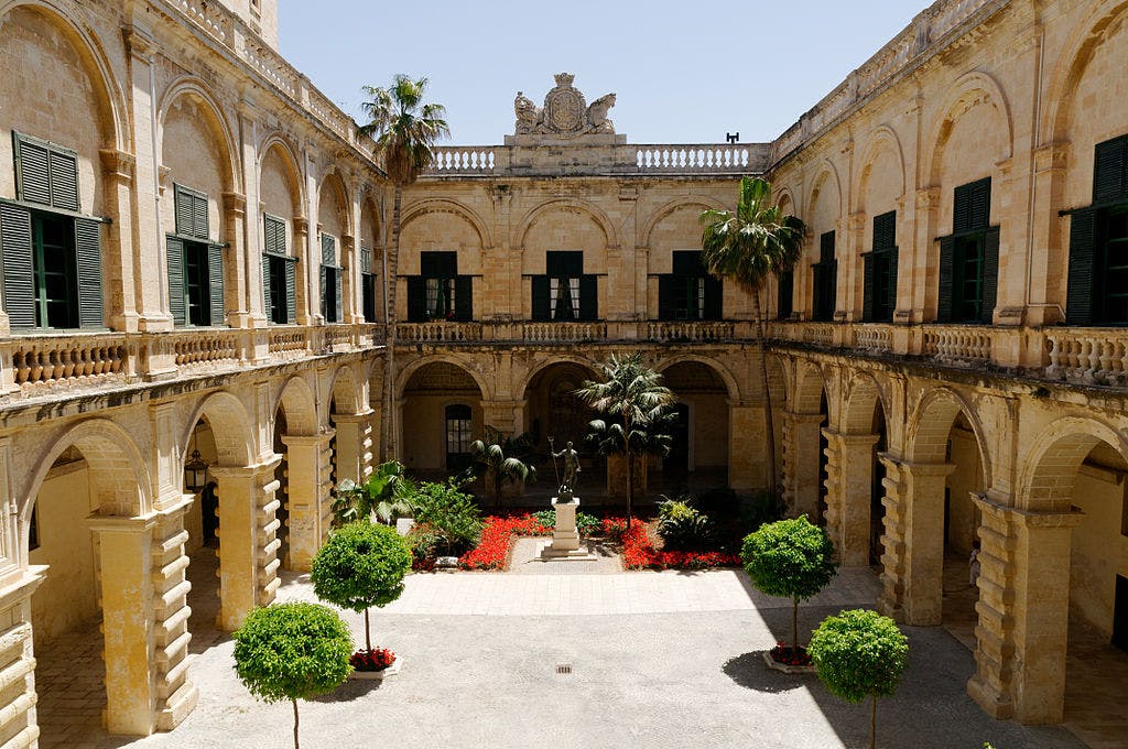 An ornate courtyard with arched colonnades, greenery, flowers, and a central statue, surrounded by beige stone buildings with green shutters.