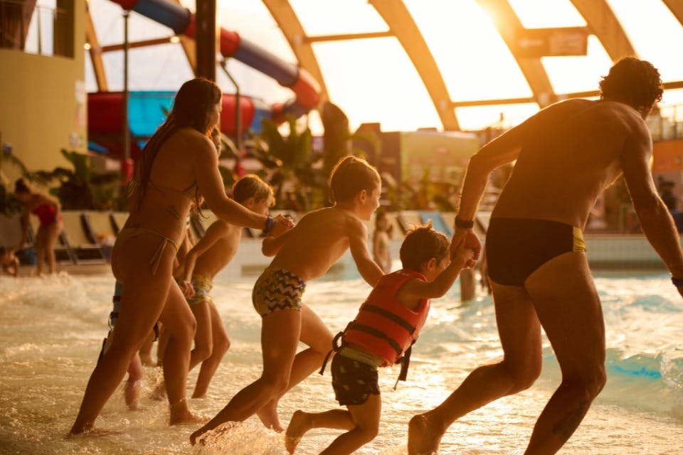 People enjoying a water park, running into a pool, with sunlight filtering through the glass roof.