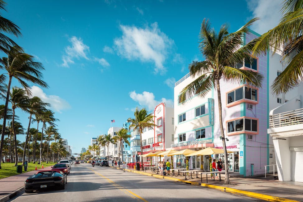 A sunny street lined with palm trees, colorful buildings, parked cars, and outdoor cafes with yellow umbrellas.