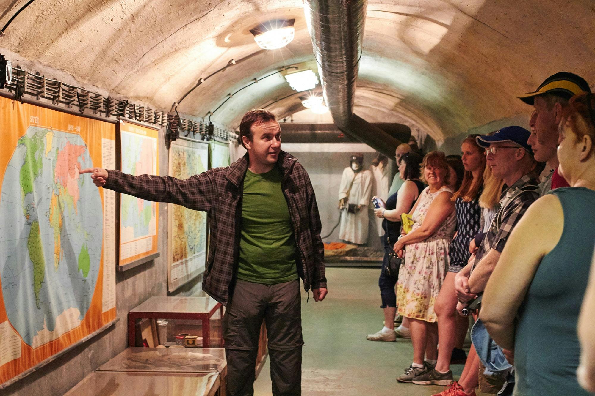 Muž v zelené košili a kostkované bundě gestikuluje na mapě na zdi uvnitř tunelu, lidé stojí a pozorují.