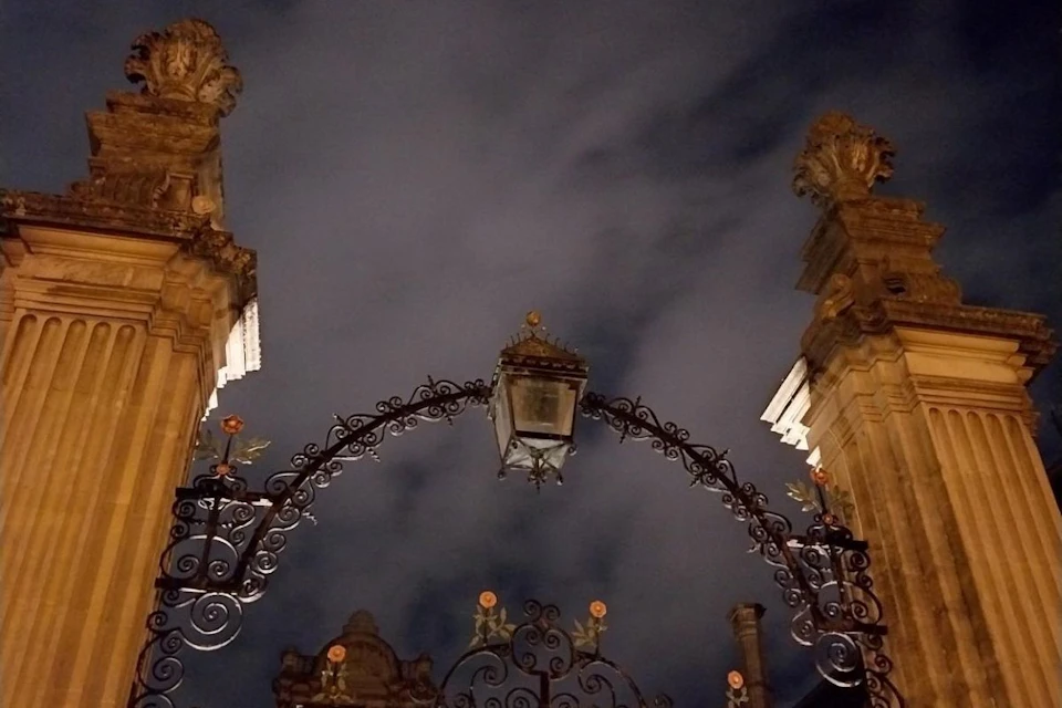 Ornate wrought iron gate with a central lantern, flanked by intricately carved stone pillars against a cloudy night sky.