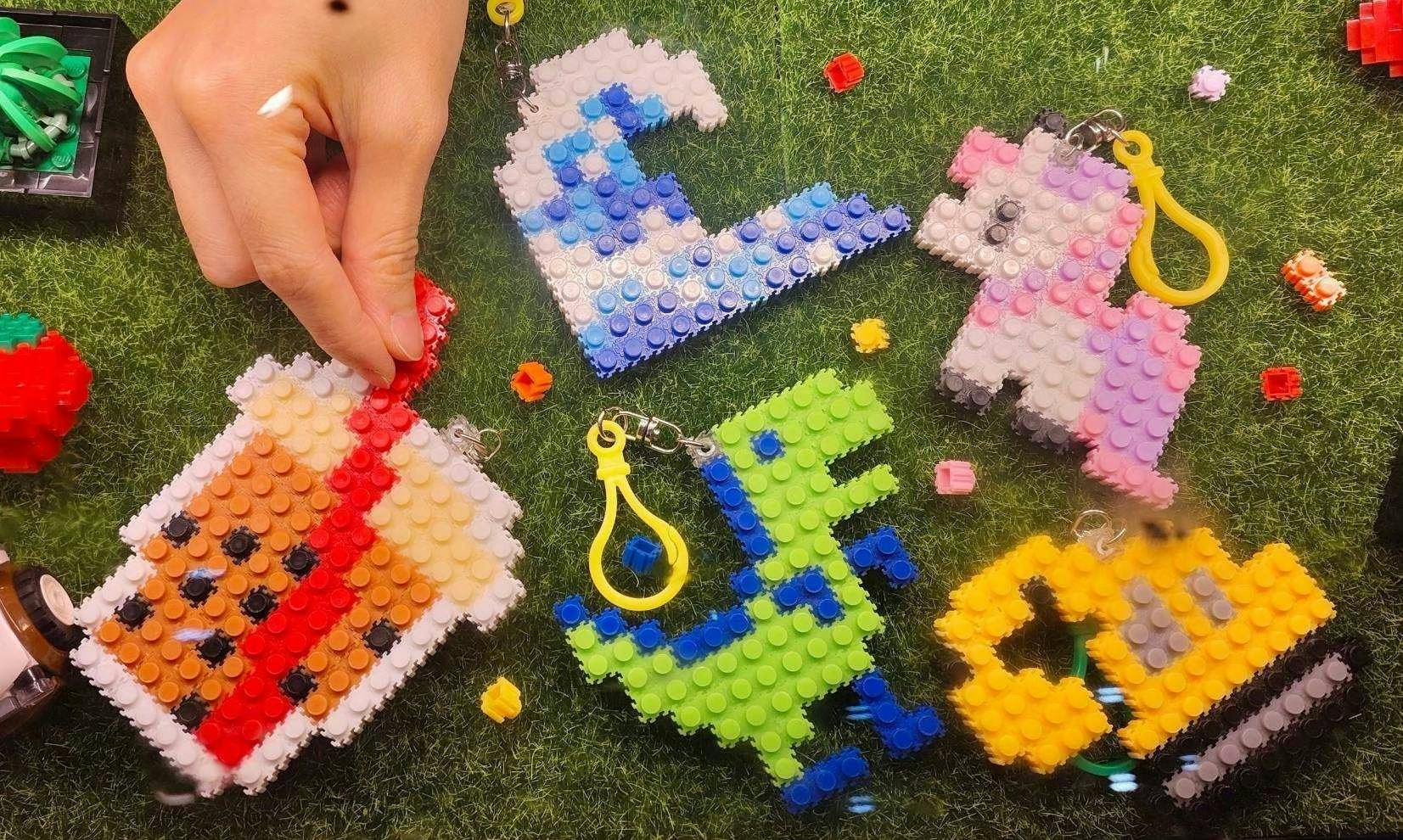 A hand arranging colorful pixelated keychains shaped like animals and objects on a grass surface.