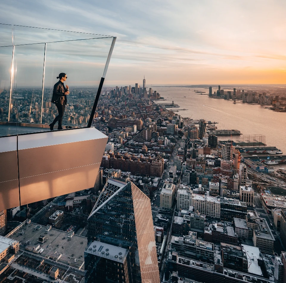 Person standing on a high glass observation deck overlooking a vast cityscape and river at sunset.