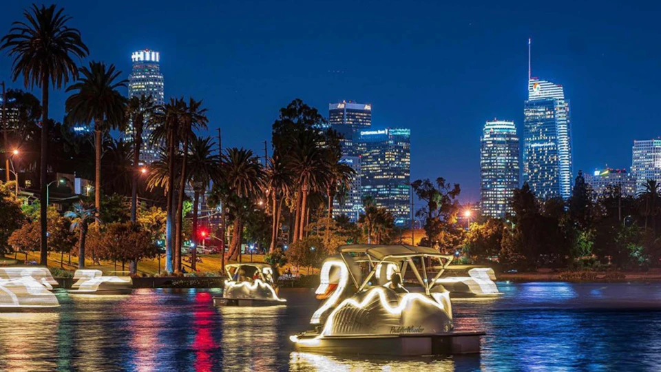 Illuminated swan boats float on a lake at night, with city skyscrapers and palm trees in the background.