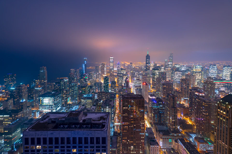 Aerial view of a brightly lit cityscape at night, featuring numerous skyscrapers and buildings with illuminated windows and streets.