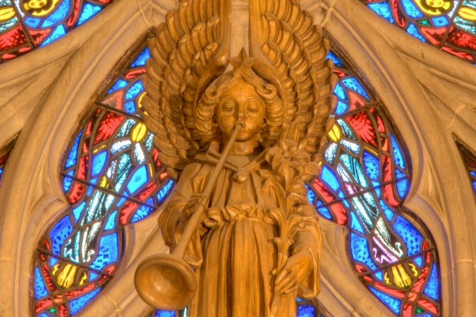 Carved wooden angel playing a trumpet, surrounded by colorful stained glass patterns in a cathedral setting.