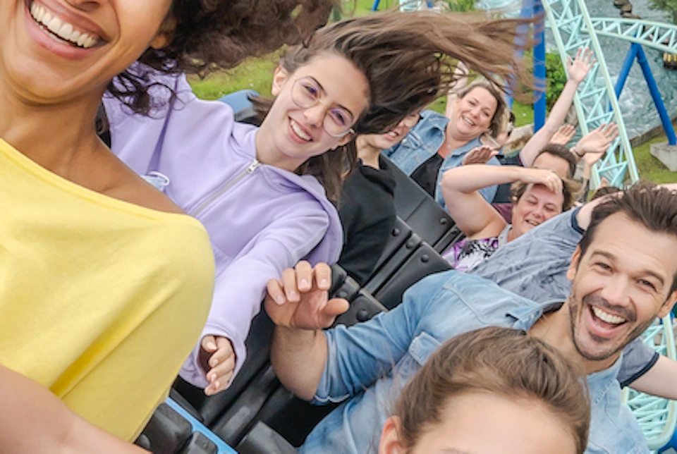People enjoying a ride on a rollercoaster, smiling and waving their arms, with a scenic view of trees and water in the background.
