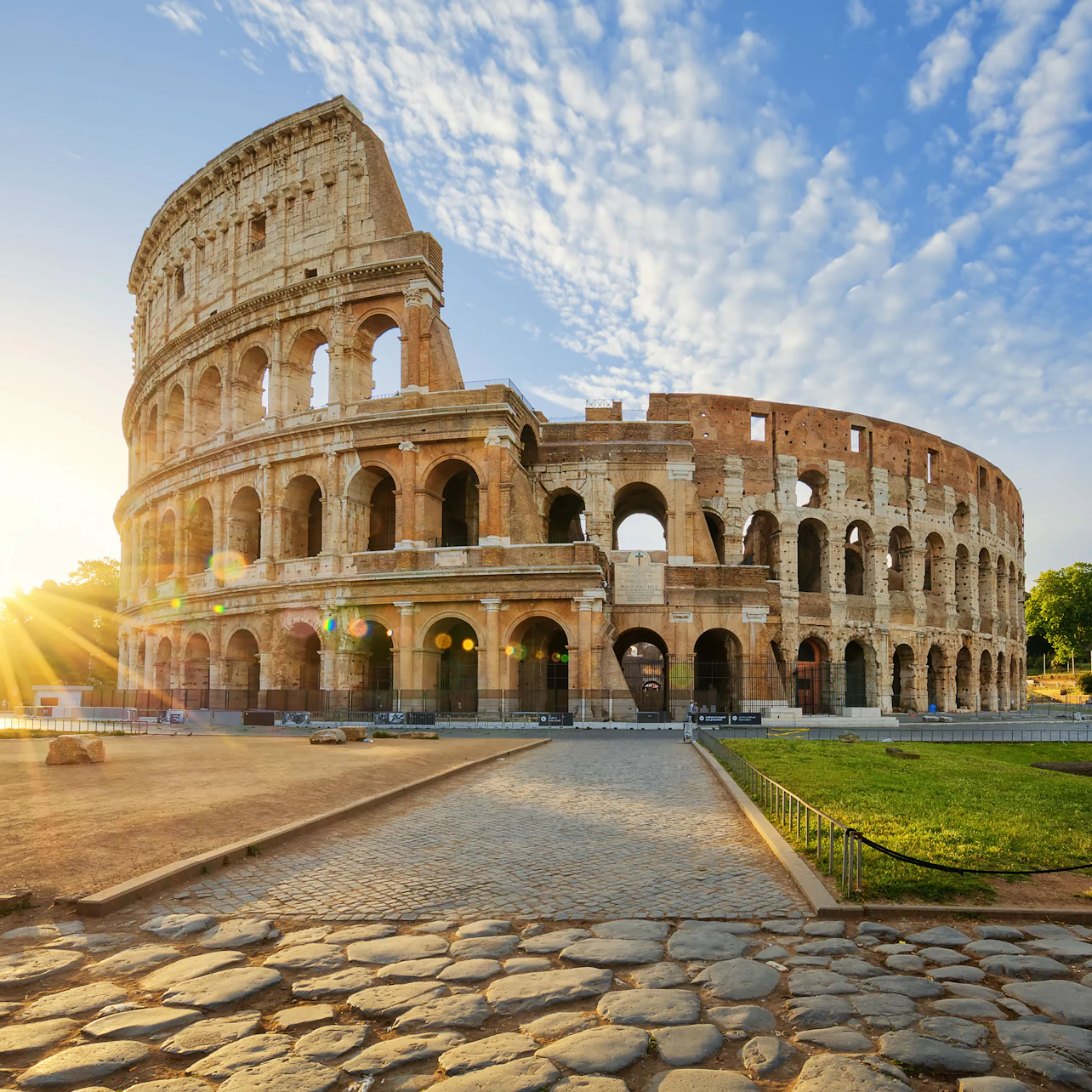 The Roman Colosseum at sunrise, with sunlight streaming through the arches and a clear sky overhead. Cobblestone path in foreground.