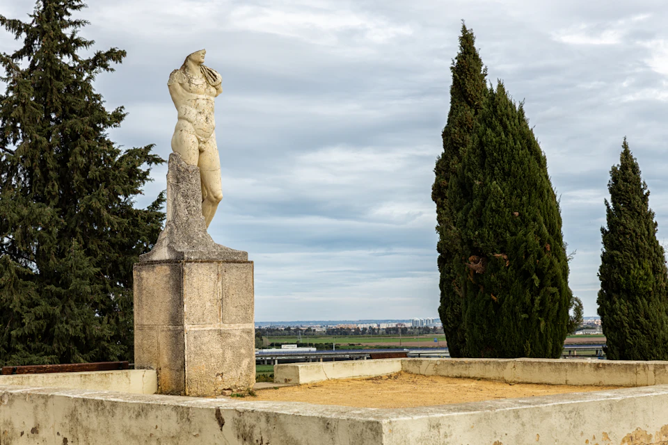 A weathered torso of a statue stands on a stone pedestal next to tall evergreen trees, against a cloudy sky and distant horizon.