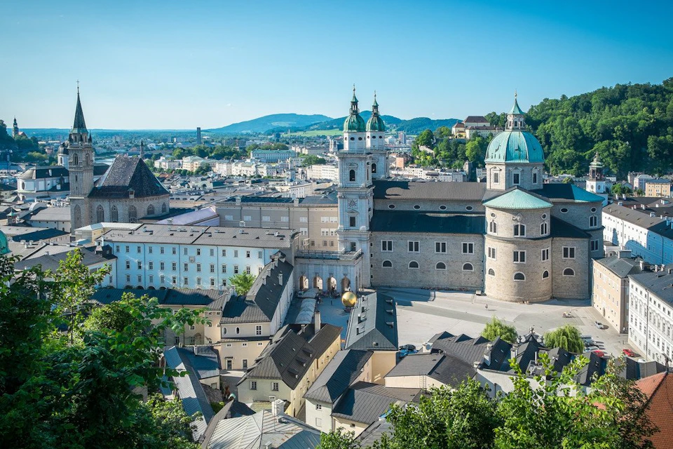 An aerial view of a city with historical buildings, featuring a cathedral with green domes, surrounded by other structures and greenery.