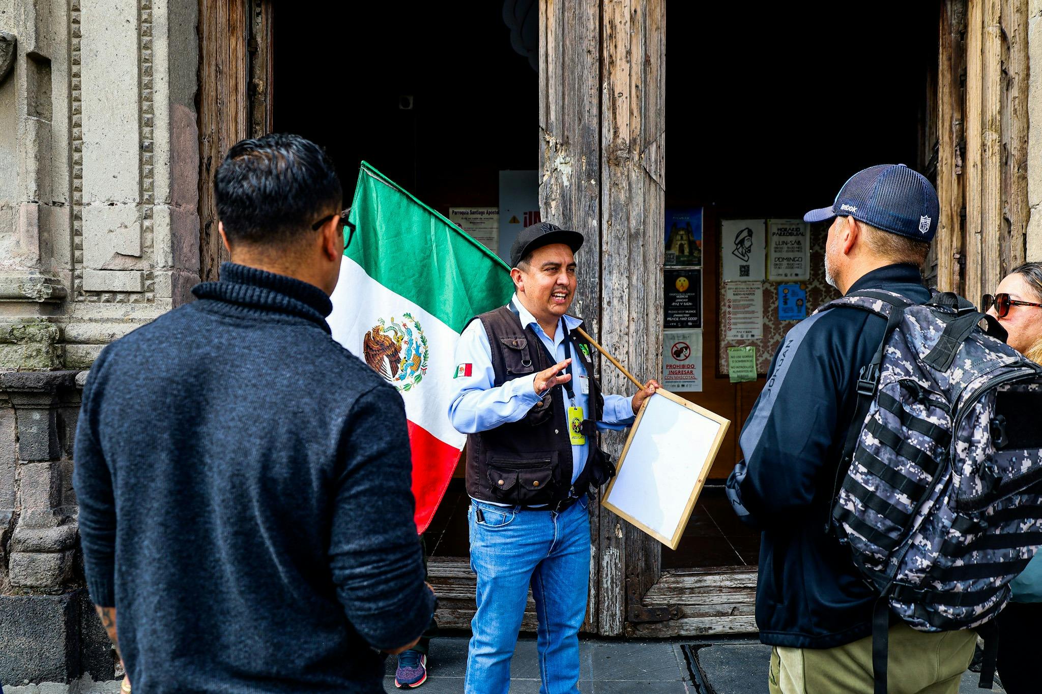 A man holding a Mexican flag and a blank whiteboard is speaking to two other people in front of a wooden doorway.