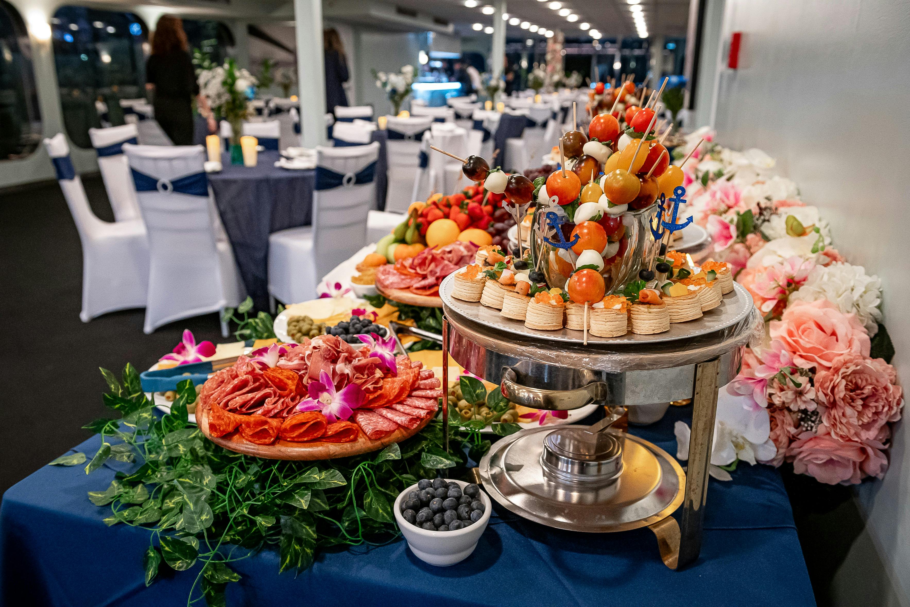 Bufetový stůl s různými předkrmy, včetně špejle, zeleniny, masa a ovoce, umístěný v hodovní síni s bílými židlemi.