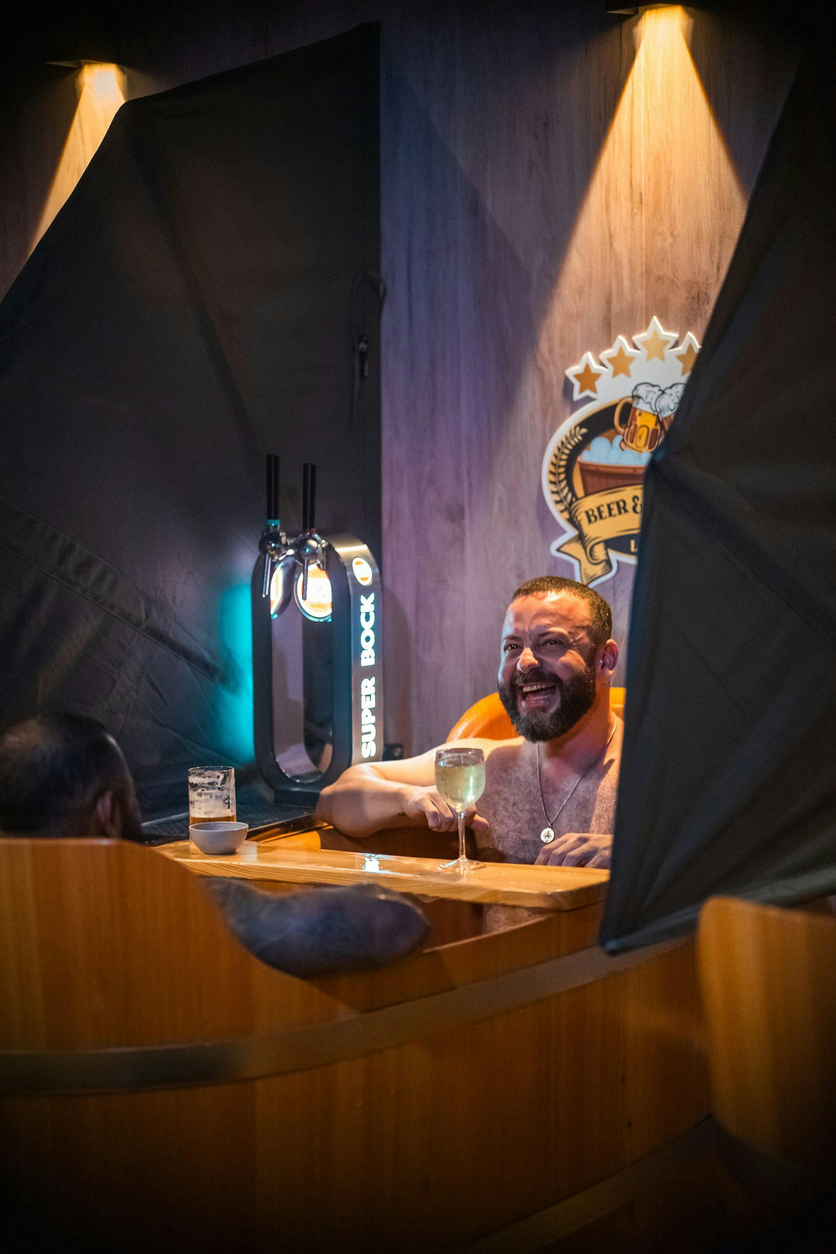 Ένας άντρας σε ένα ξύλινο τζακούζι χαμογελάει κρατώντας ένα ποτήρι κρασιού, με βρύσες μπύρας και το λογότυπο "Beer Spa" στον τοίχο πίσω του.