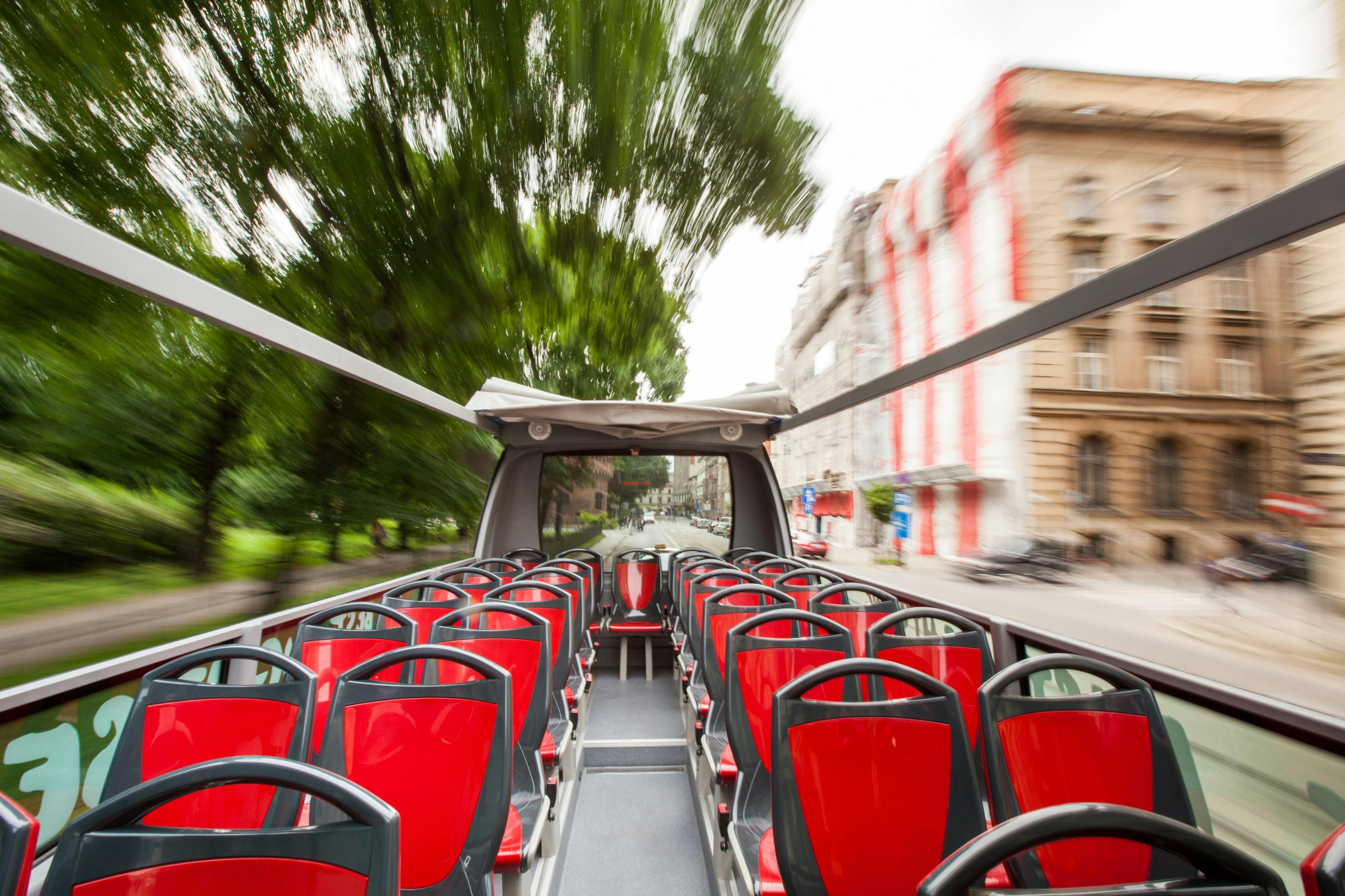 Empty red and black seats on the upper deck of a moving open-top bus passing through a city street with blurred buildings and trees.