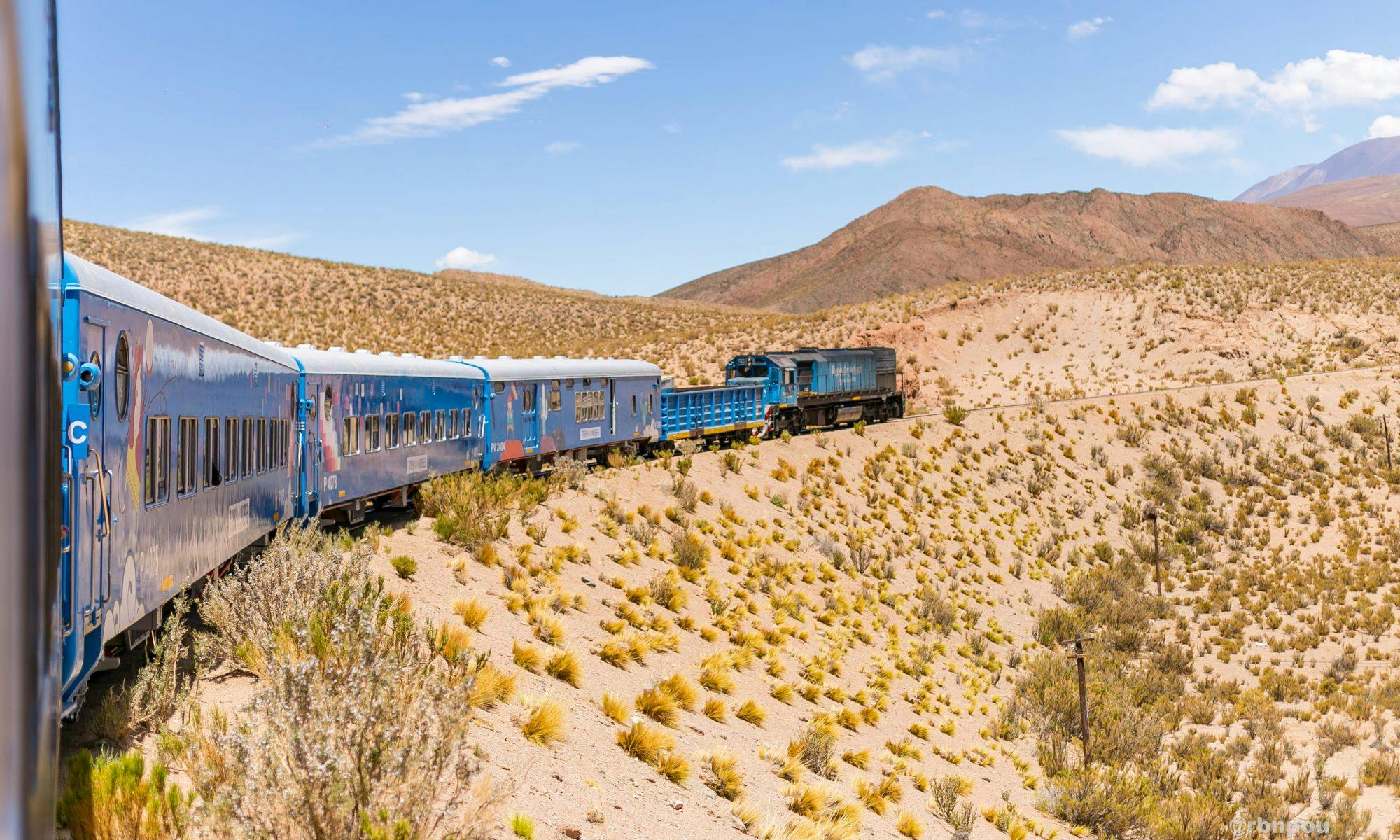 A blue train travels through a desert landscape with dry vegetation and distant mountains under a clear blue sky.