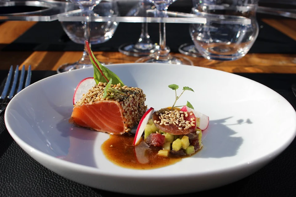 A plate of sesame-crusted salmon served with sauce, garnish and a small selection of vegetables, arranged on a white plate with wine glasses in the background.