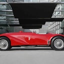 Classica auto sportiva decappottabile rossa parcheggiata in un ambiente urbano moderno con pareti di vetro e grandi pannelli neri sopra la testa.