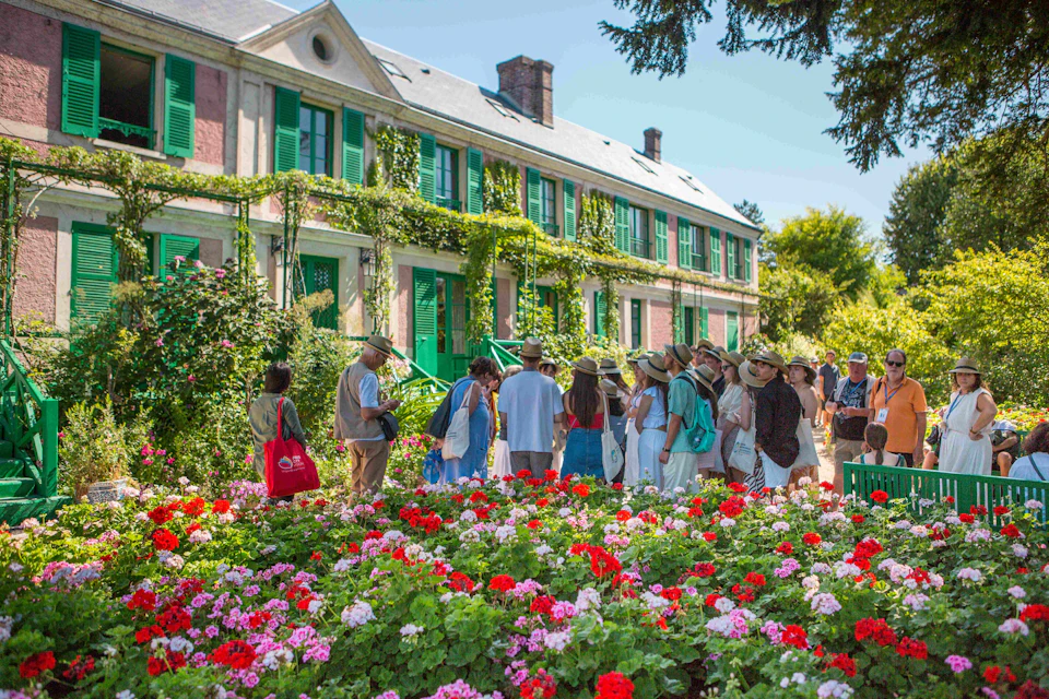 Group of people in summer attire tour a flower garden with vibrant blooms in front of a vine-covered, green-shuttered house.