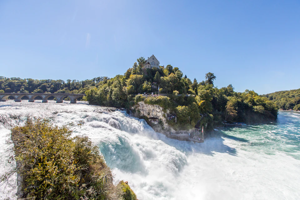 Large waterfall cascading down rocks with a tree-covered hill and a castle-like building on top, under a clear blue sky.