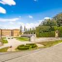 Historic palace with manicured gardens, hedges, statues, and pathways under a blue sky with scattered clouds.