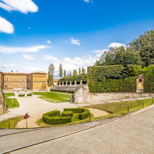 Historic palace with manicured gardens, hedges, statues, and pathways under a blue sky with scattered clouds.
