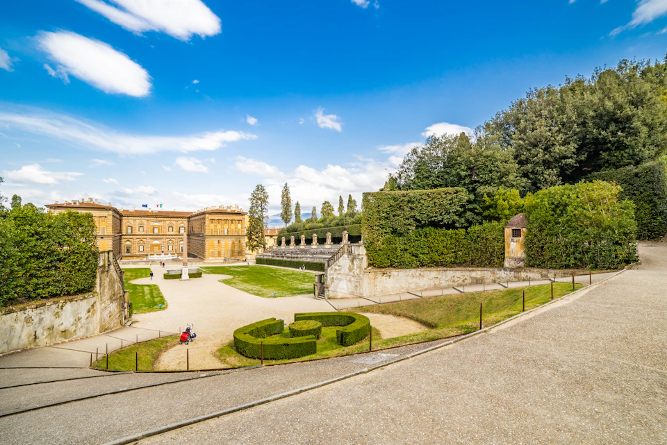 Historic palace with manicured gardens, hedges, statues, and pathways under a blue sky with scattered clouds.