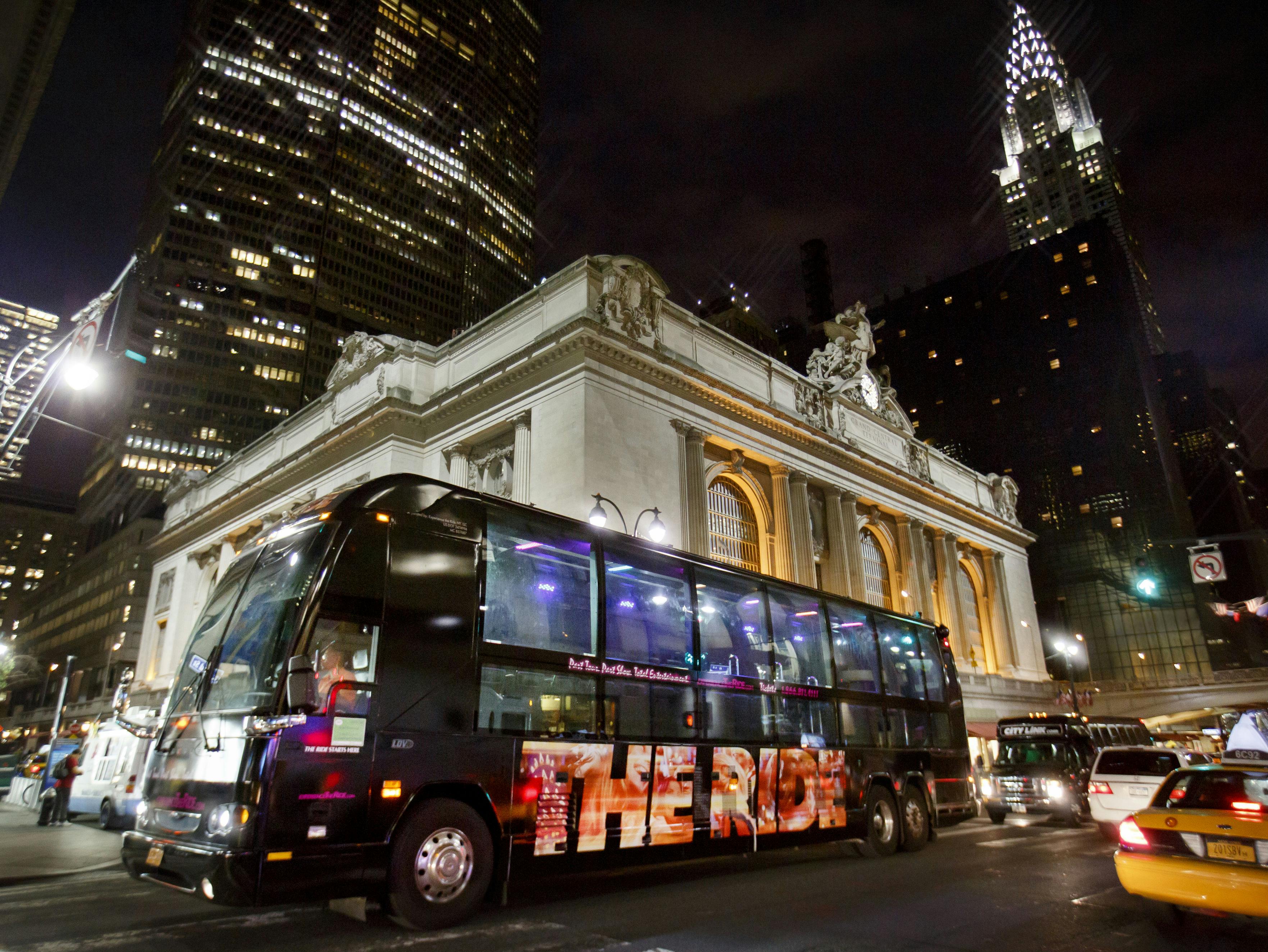 Ένα τουριστικό λεωφορείο με την ένδειξη «Η ΒΟΛΤΑ» περνάει μπροστά από ένα μεγαλοπρεπές, φωτισμένο κτίριο τη νύχτα σε μια πόλη με ψηλούς ουρανοξύστες.