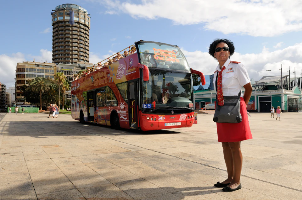 A tour guide stands in front of a red double-decker bus parked in an open area on a sunny day.