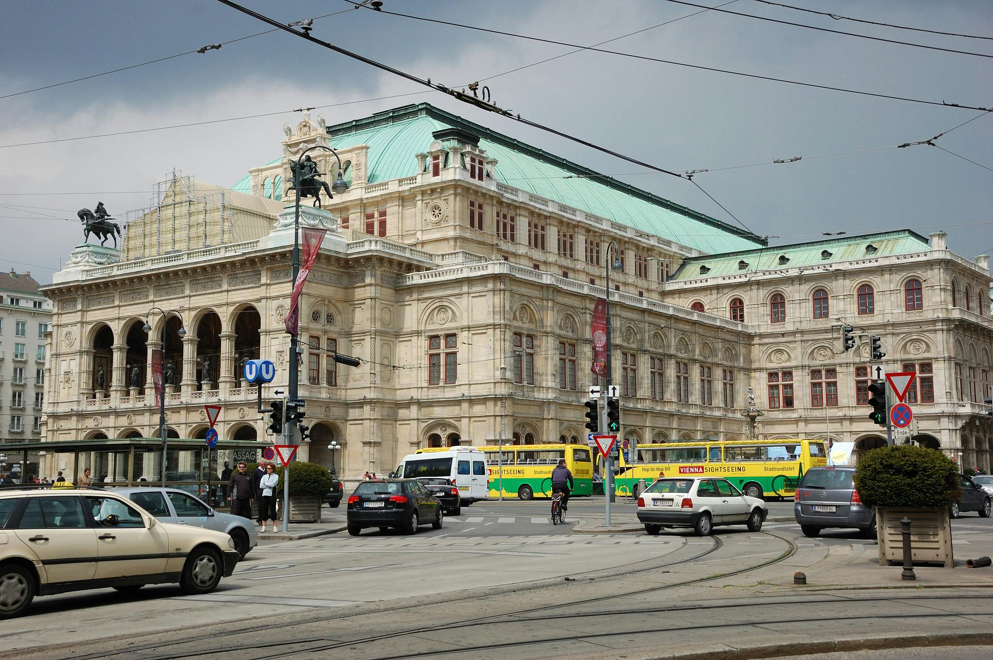 Historisk beige byggnad med grönt tak, statyer och välvda fönster. Bilar, bussar och cyklister i korsningen; spårvagnsspår i förgrunden.