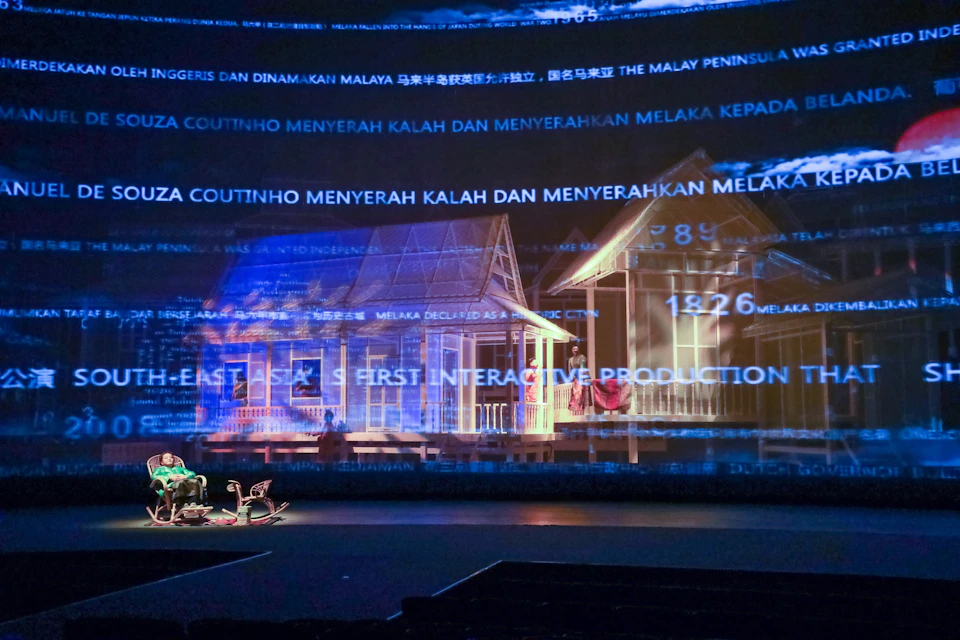 Stage with a digital projection featuring text, a traditional house, and performers seated at the front.