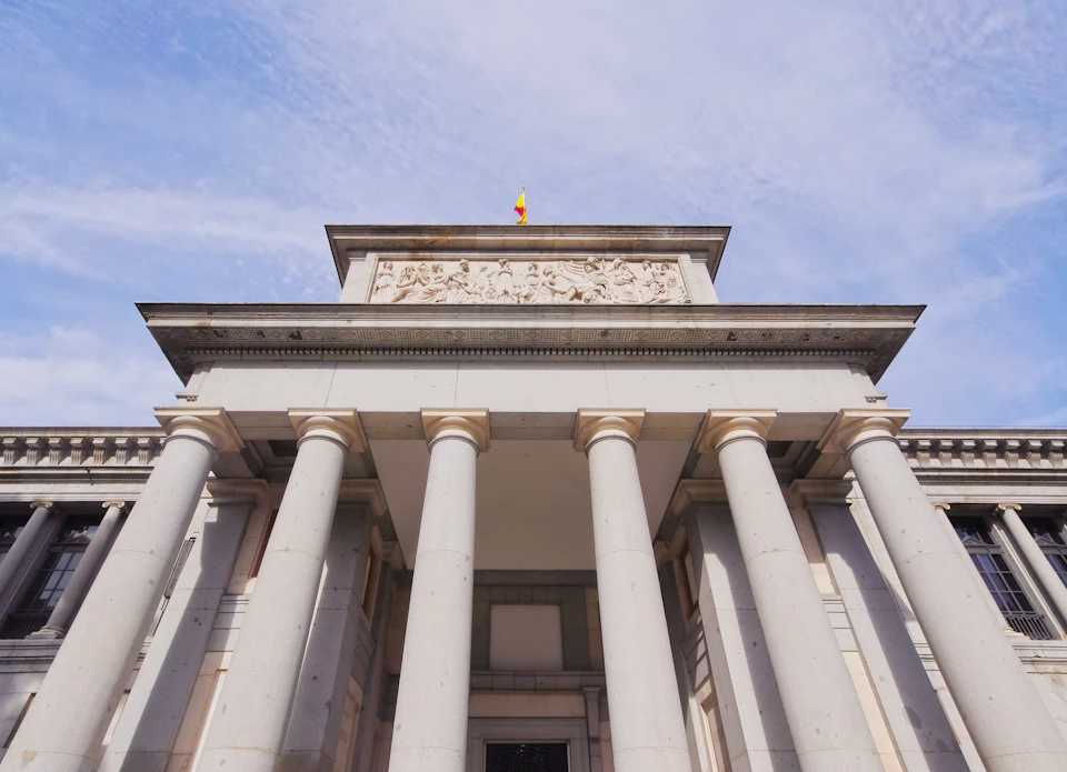 Front view of a neoclassical building with tall, gray columns, a detailed frieze, and a small flag on top, all under a partly cloudy sky.