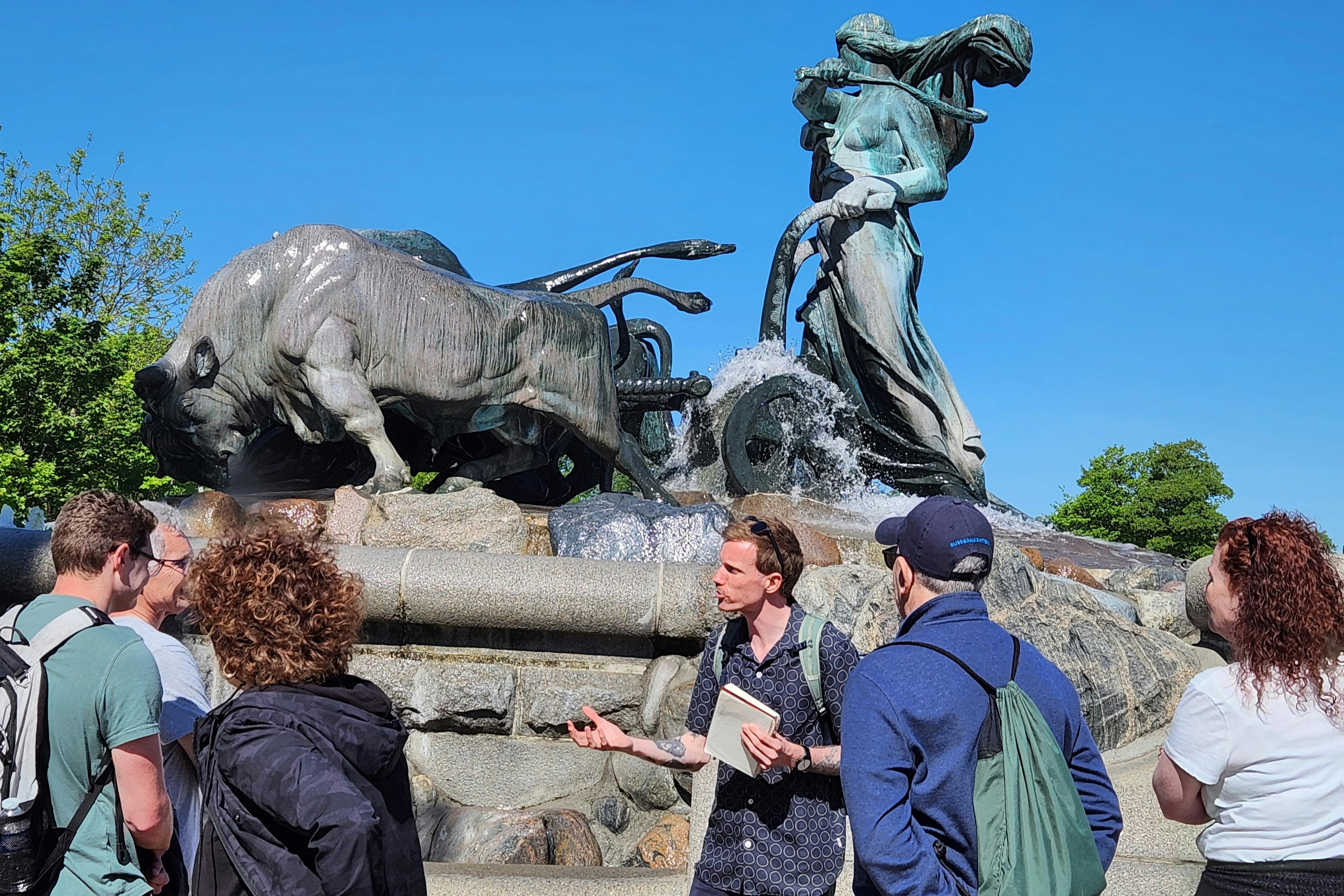 A tour guide with a notebook gestures while speaking to a group near a large fountain with statues of oxen and a woman.
