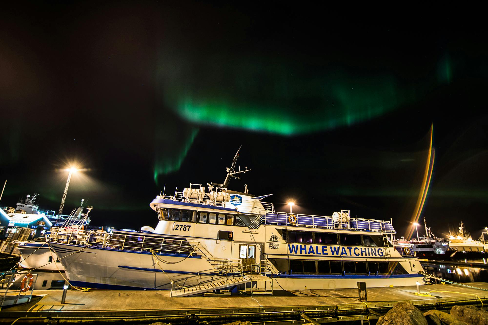 A whale-watching tour boat docked at night, with green Northern Lights visible in the sky above.