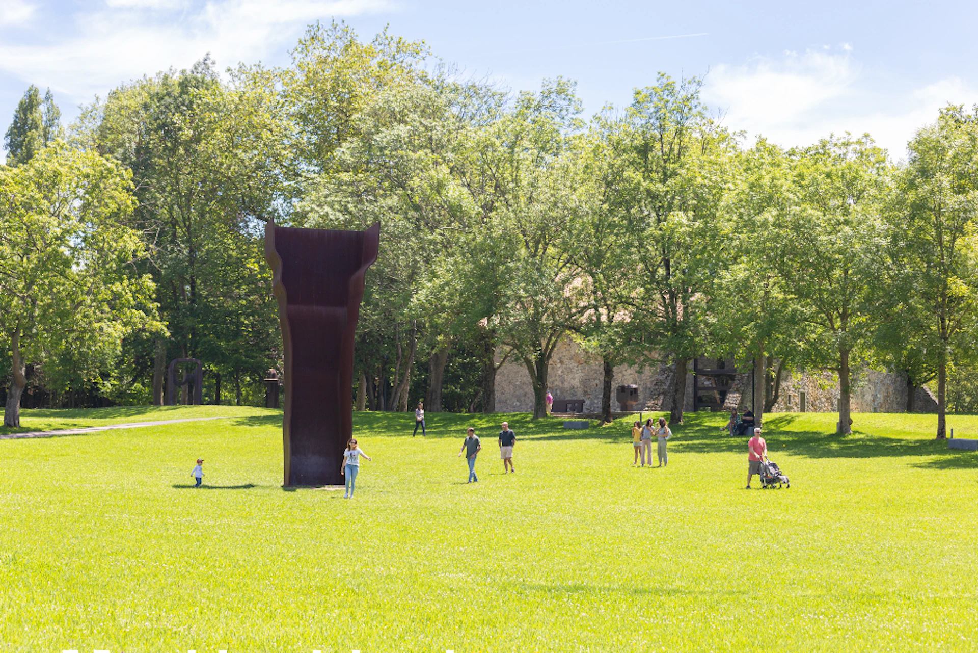 People walking and standing near a large outdoor sculpture in a sunny park with green grass and trees.