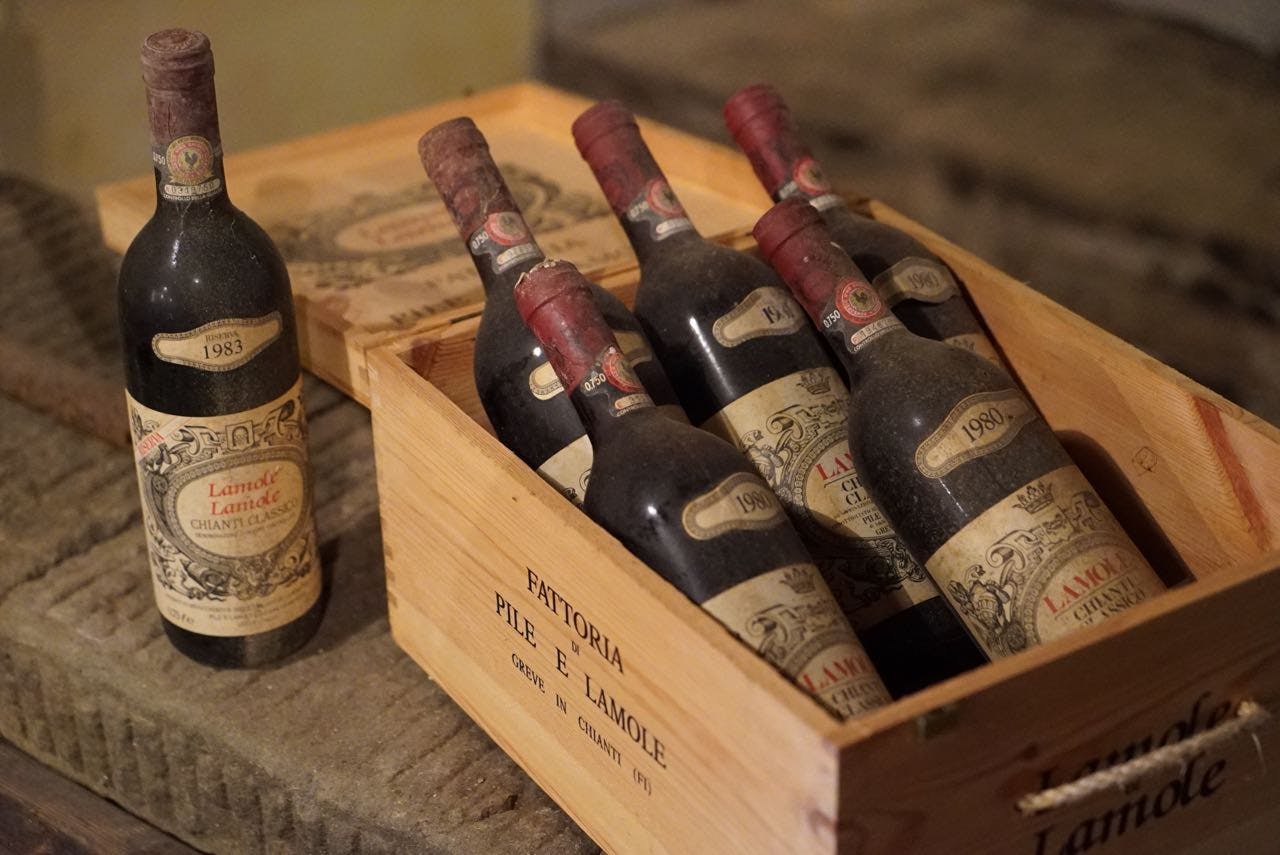 Έξι σκονισμένα μπουκάλια κρασιού σε ένα ξύλινο καφάσι με την ένδειξη "Fattoria Pile e Lamole", με ένα όρθιο μπουκάλι δίπλα του.