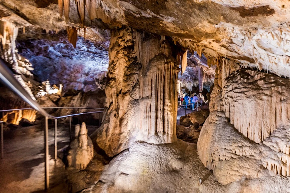 A group of people is walking through a cavern with large stalactites and stalagmites, illuminated by artificial lighting.