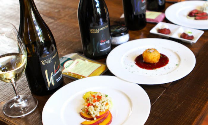 Dvě jídla, jedno s kuskusem a broskvovými plátky, druhé se smaženým předmětem na omáčce, vedle lahví s vínem s označením „Williamson Wines“.