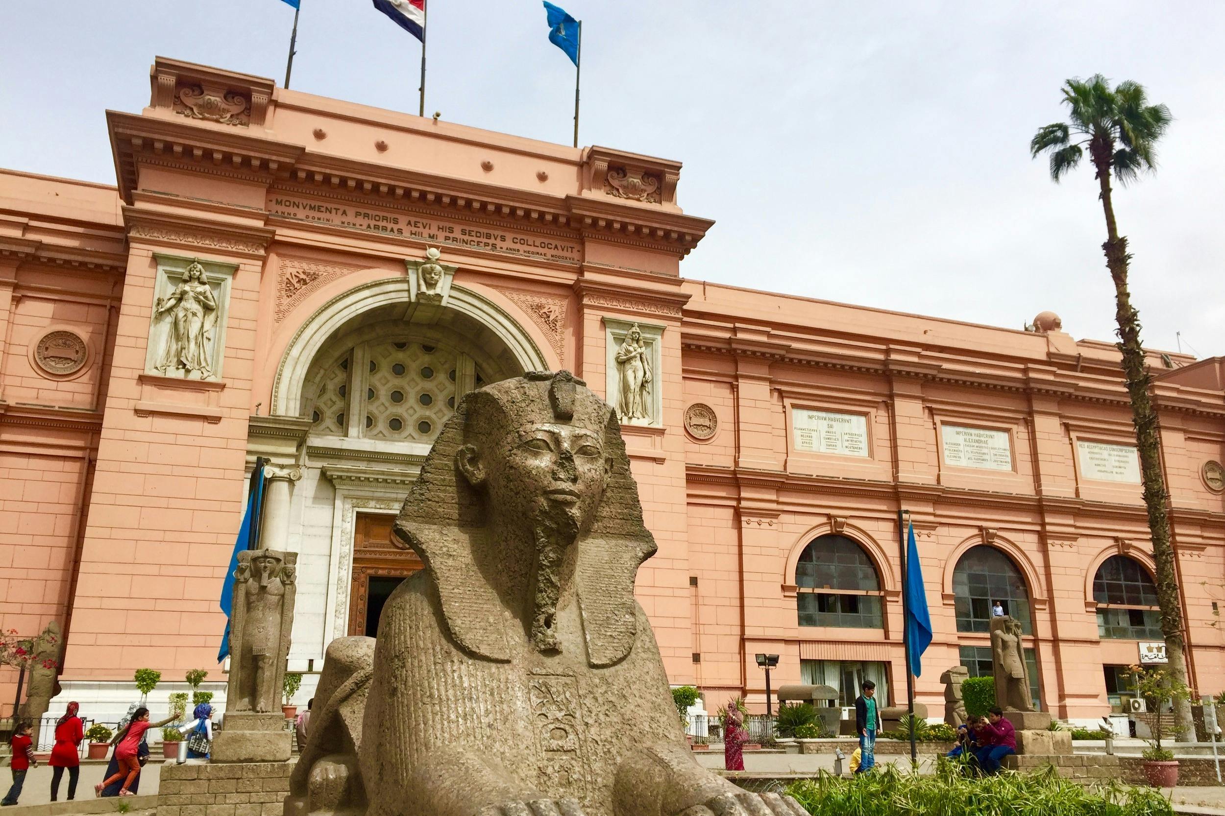 A statue of a sphinx in front of a pink building with arched windows and flags on the roof, surrounded by people and greenery.